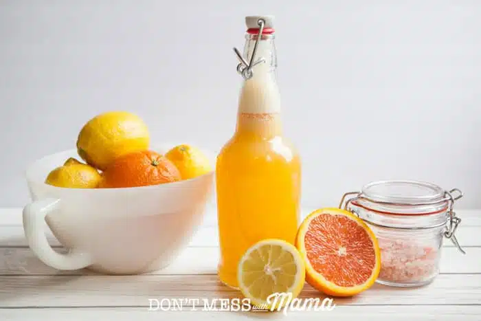 A side shot of a homemade electrolyte drink in a glass bottle on a white surface with citrus fruits