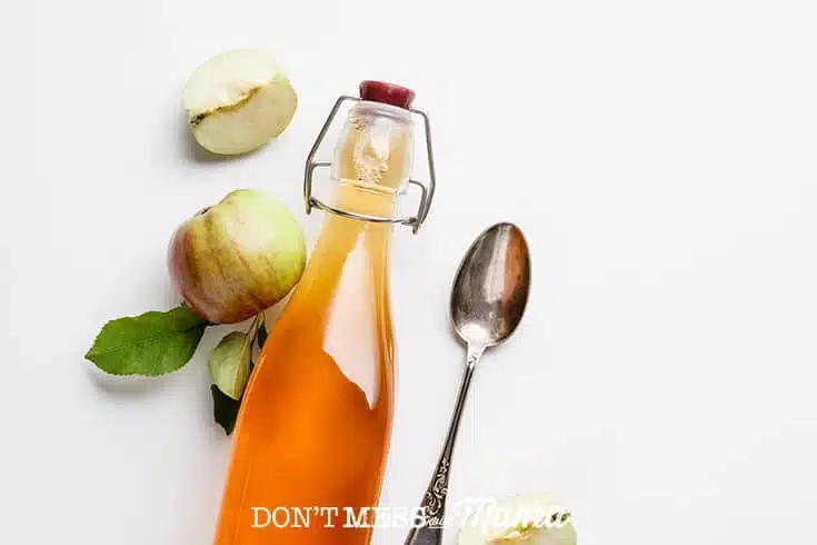 Closeup of apple cider vinegar in a glass bottle on a table with a spoon and apples