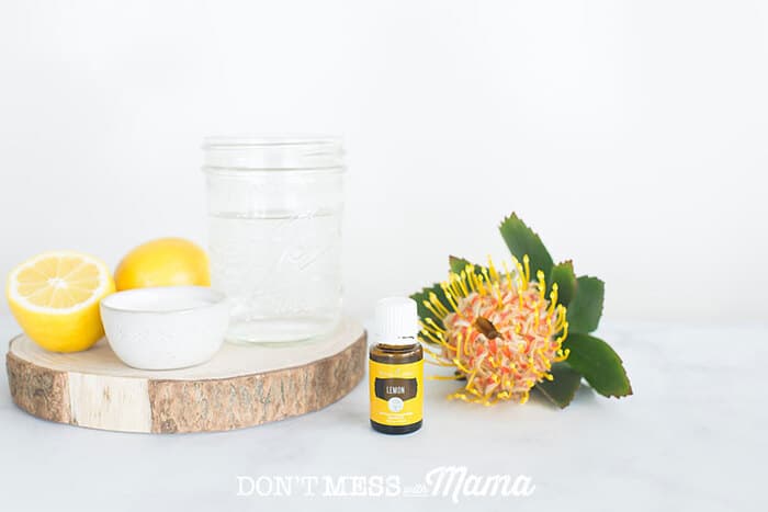 Closeup of lemon essential oil on a table with a flower and cup of water in the background