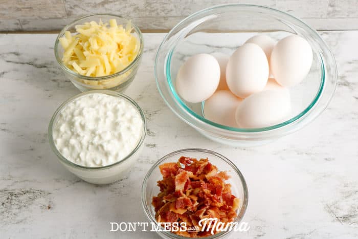 Bowl of eggs, bowl of cottage cheese, bowl of bacon, bowl of shredded gruyere cheese on a white table