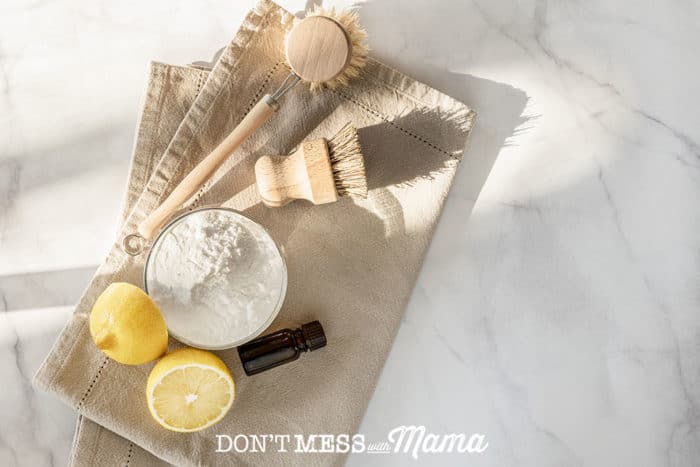 baking soda in a bowl, essential oils, lemon cut in half, and cleaning brushes on a brown towel on a countertop