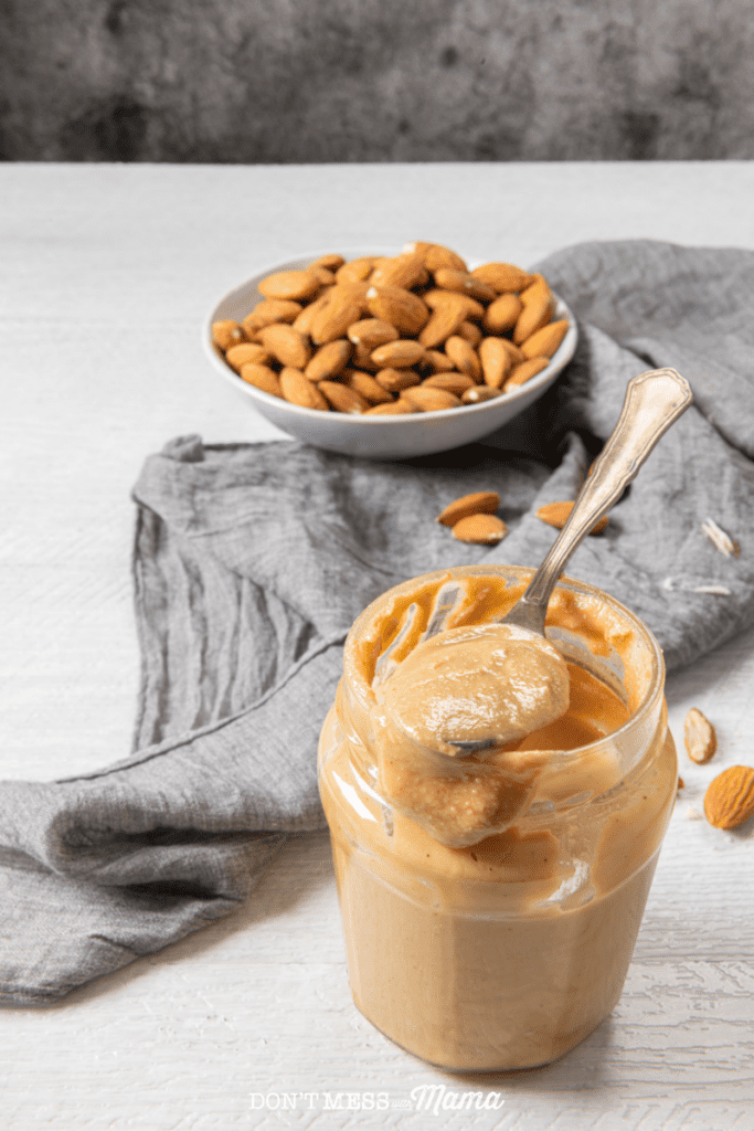 creamy almond butter on a spoon on top of a glass jar with a gray towel and bowl of almonds in the background