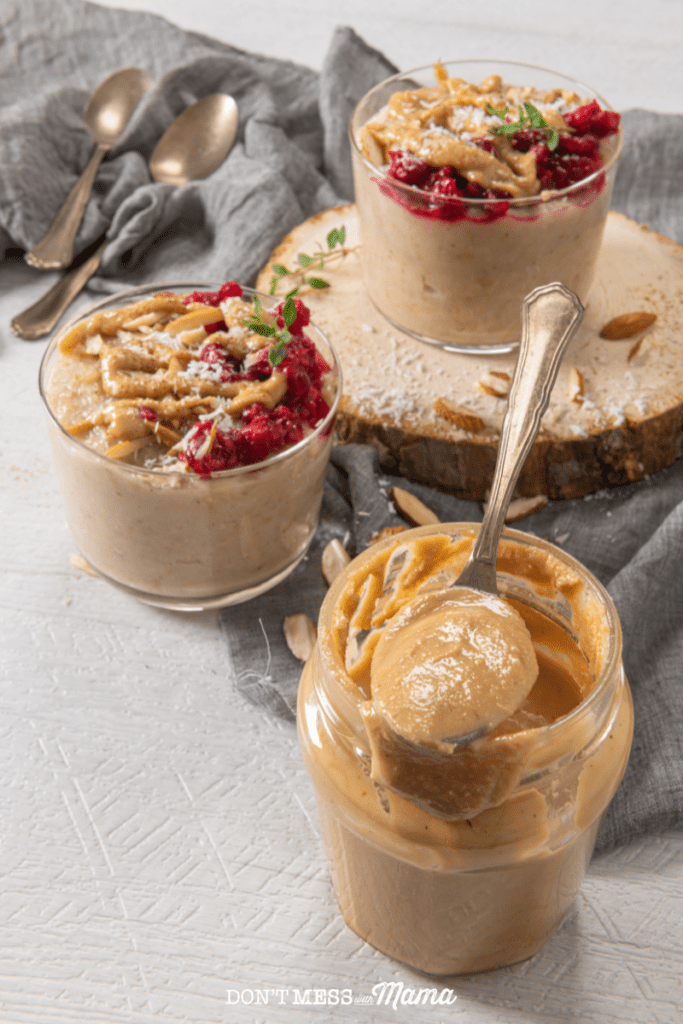 almond butter on a spoon on top of a glass jar filled with almond butter with 2 glass cups of oats drizzled with almond butter nearby on a table