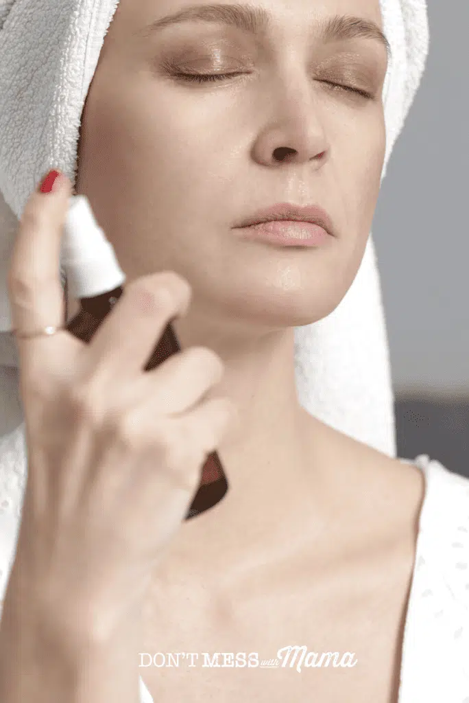 woman with white towel on head spraying facial spray