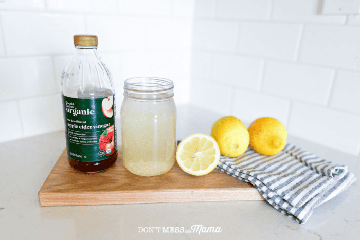 bottle of apple cider vinegar, lemons, and water on a cutting board on a kitchen counter