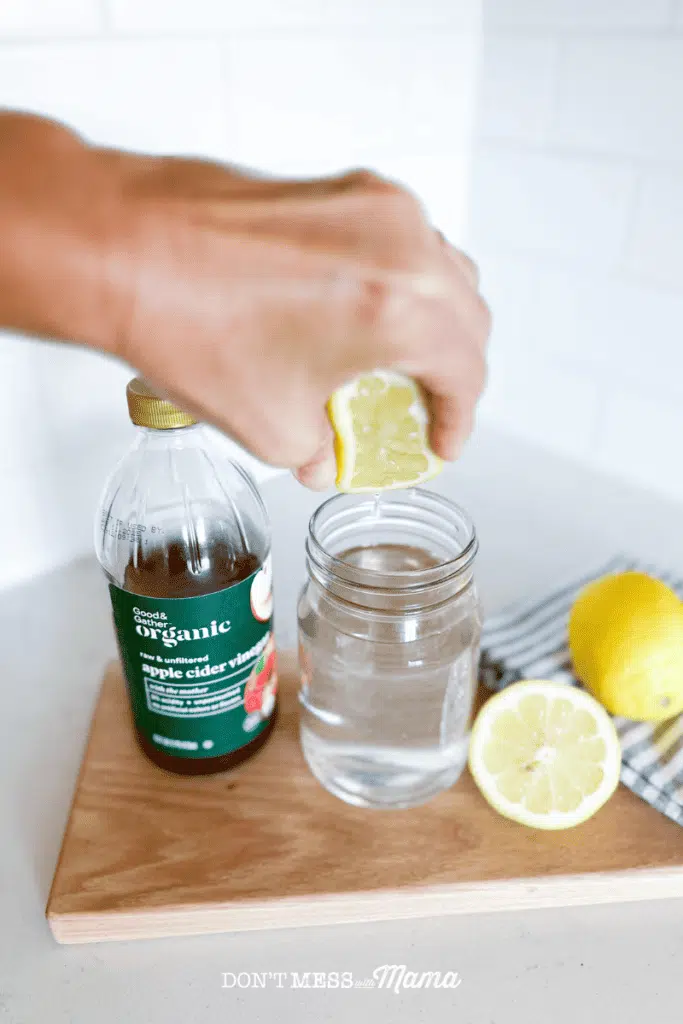 squeezing lemon juice into a glass of water with a bottle of apple cider vinegar and lemons on a cutting board 