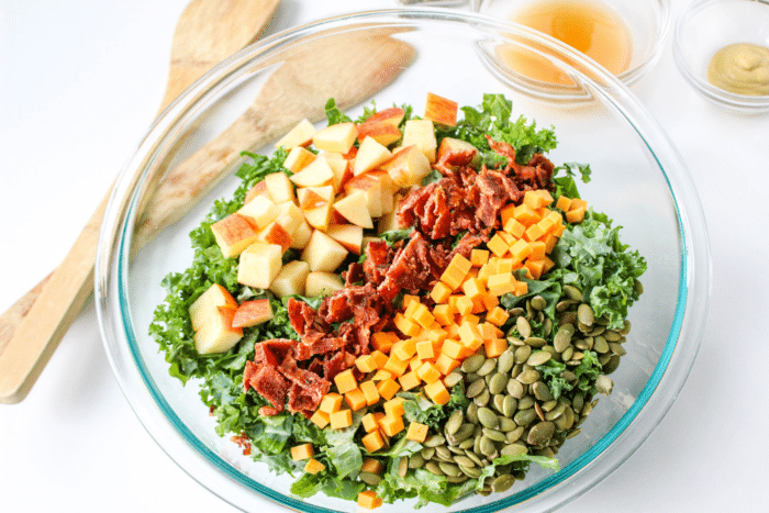 clear glass bowl with kale salad and apples, bacon, cheese, pepitas layered on top